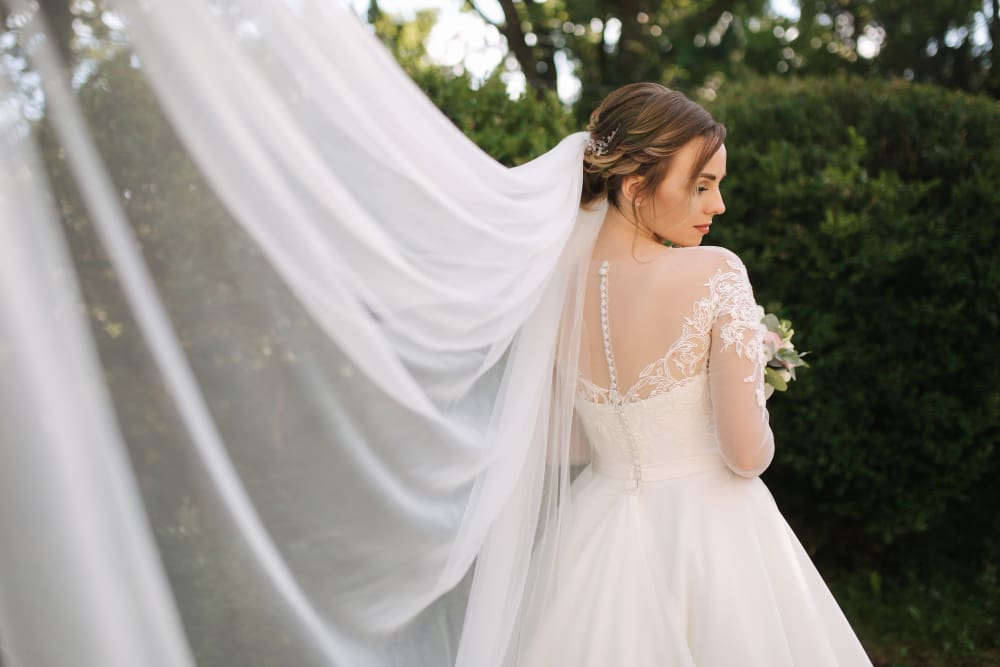A bride in a long-sleeved lace wedding dress stands outdoors, looking over her shoulder, with her veil flowing behind her.