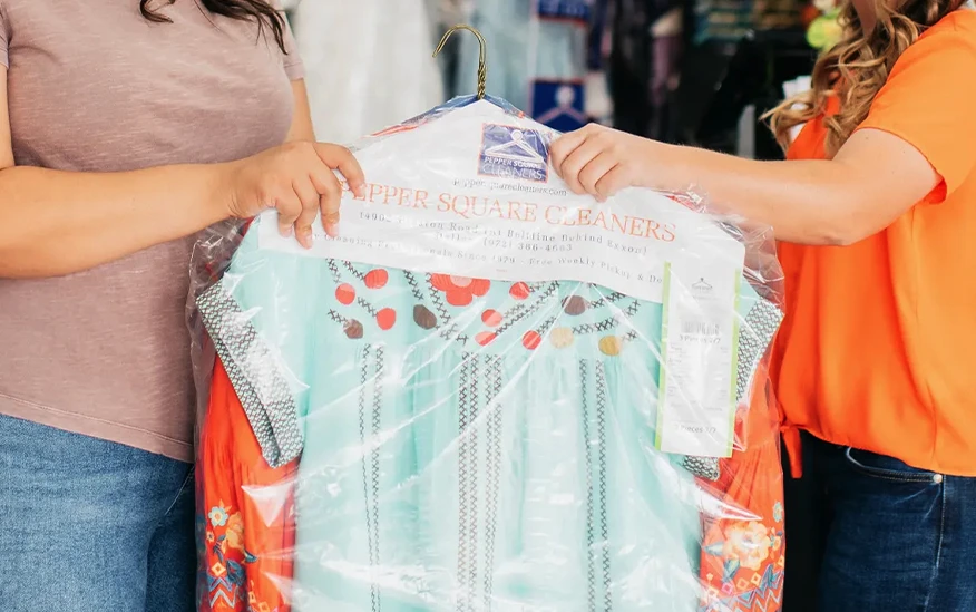Two people hold a dress covered in clear plastic from a dry cleaner, with only their torsos and arms visible.