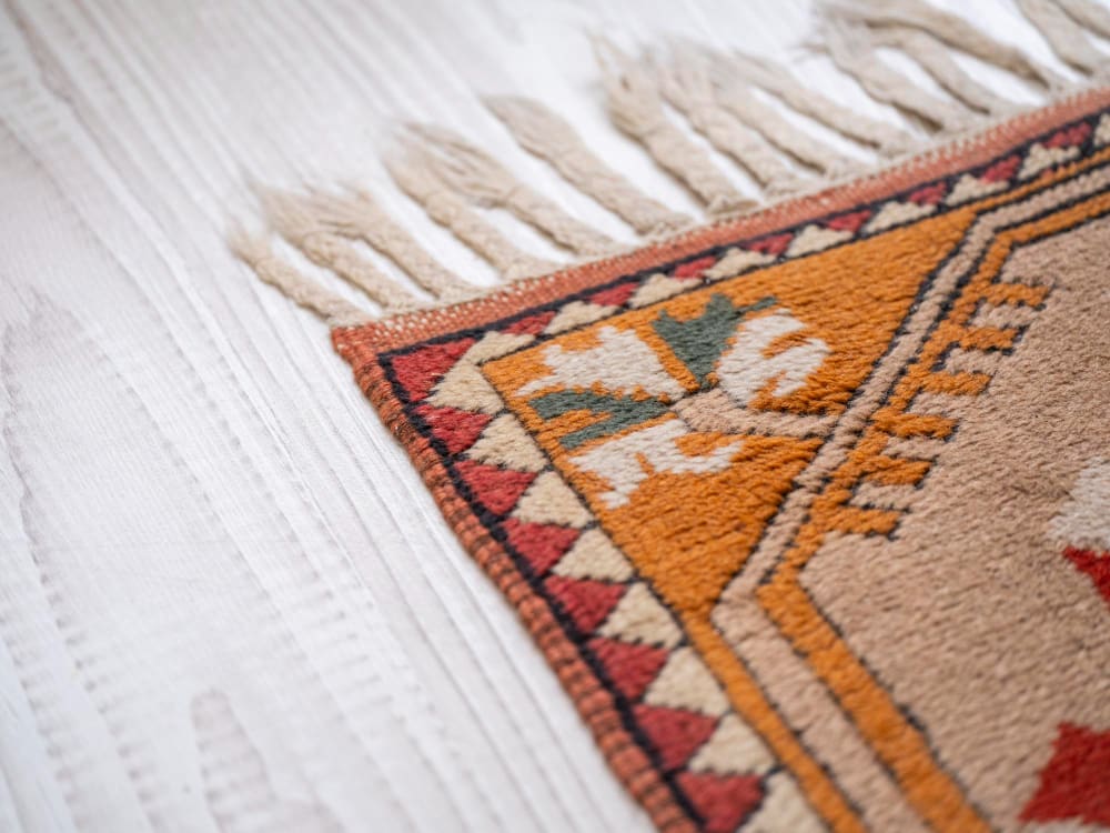 Close-up of a decorative rug with a floral pattern, fringed edge, and geometric border, lying on a light-colored wooden floor.