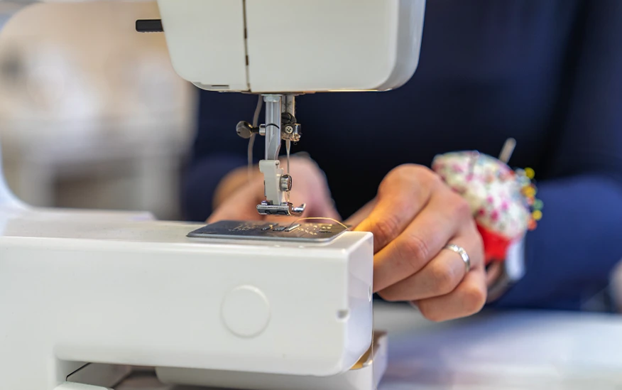 Close-up of a person using a sewing machine, guiding fabric under the needle, with a pincushion worn on their hand.