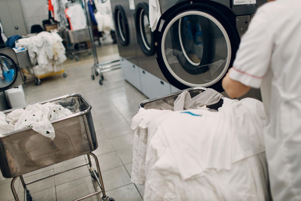 A worker loads white laundry into an industrial washing machine in a laundry facility, with metal carts holding more laundry nearby.