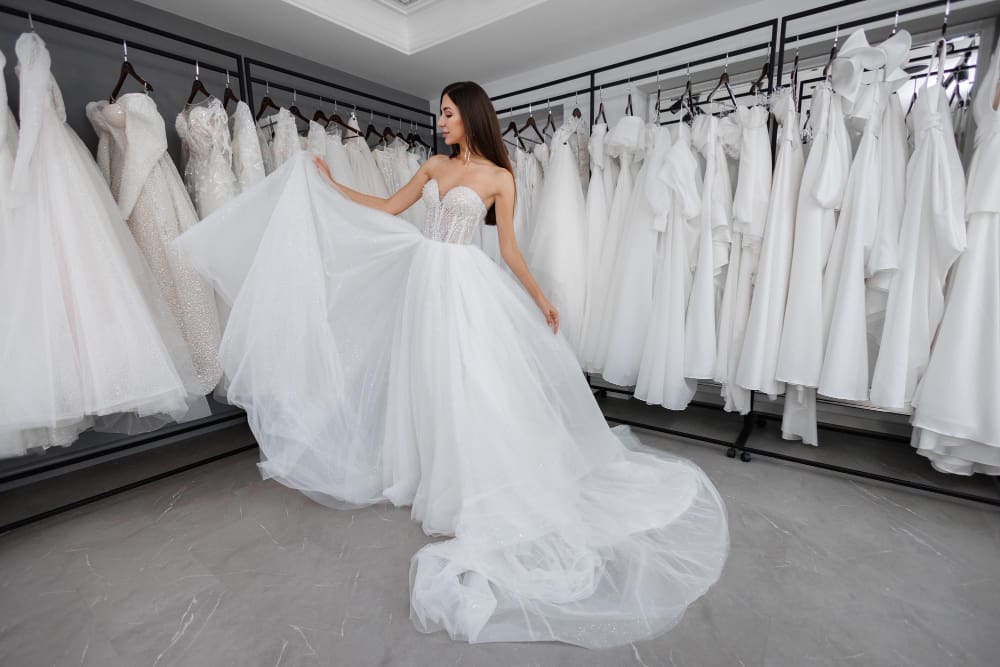A woman in a strapless white wedding gown stands in a bridal boutique, surrounded by rows of white dresses on racks.