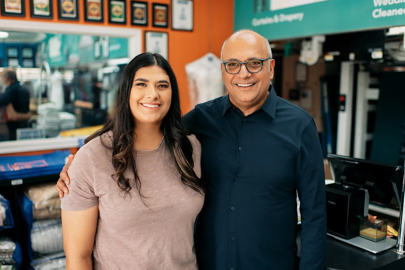 Two people stand side by side, smiling, inside a business with shelves and framed certificates visible in the background.