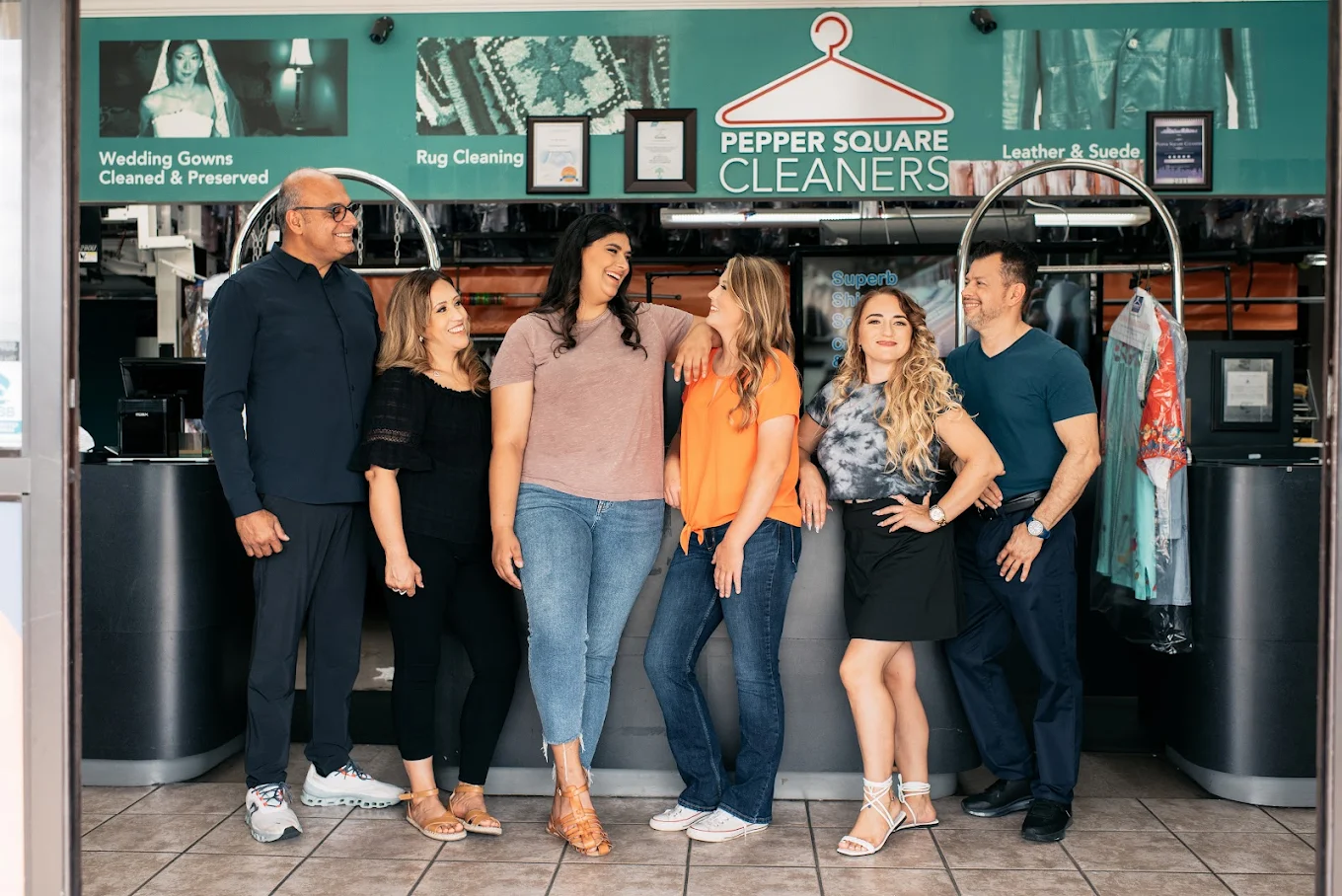 Six people stand in front of the counter at Pepper Square Cleaners, posing and smiling for a group photo inside the dry cleaning shop.