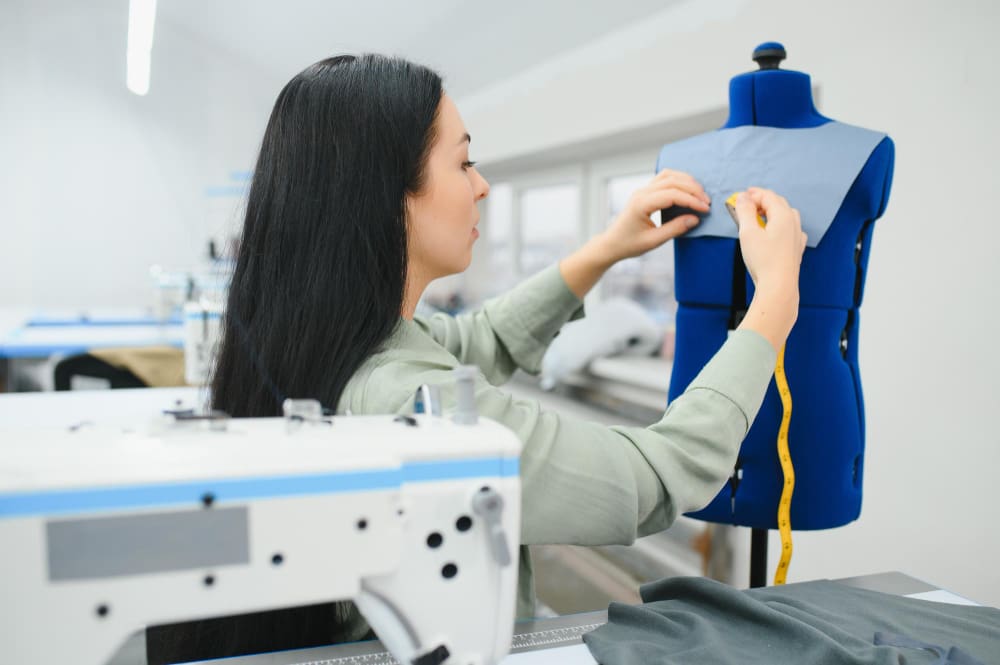 A woman is measuring and pinning blue fabric on a dress form in a sewing studio, with a sewing machine visible in the foreground.