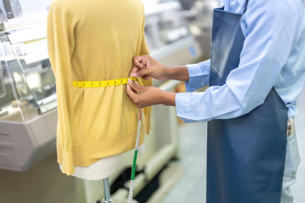 A person wearing an apron uses a measuring tape to take the waist measurement of a mannequin dressed in a yellow sweater.