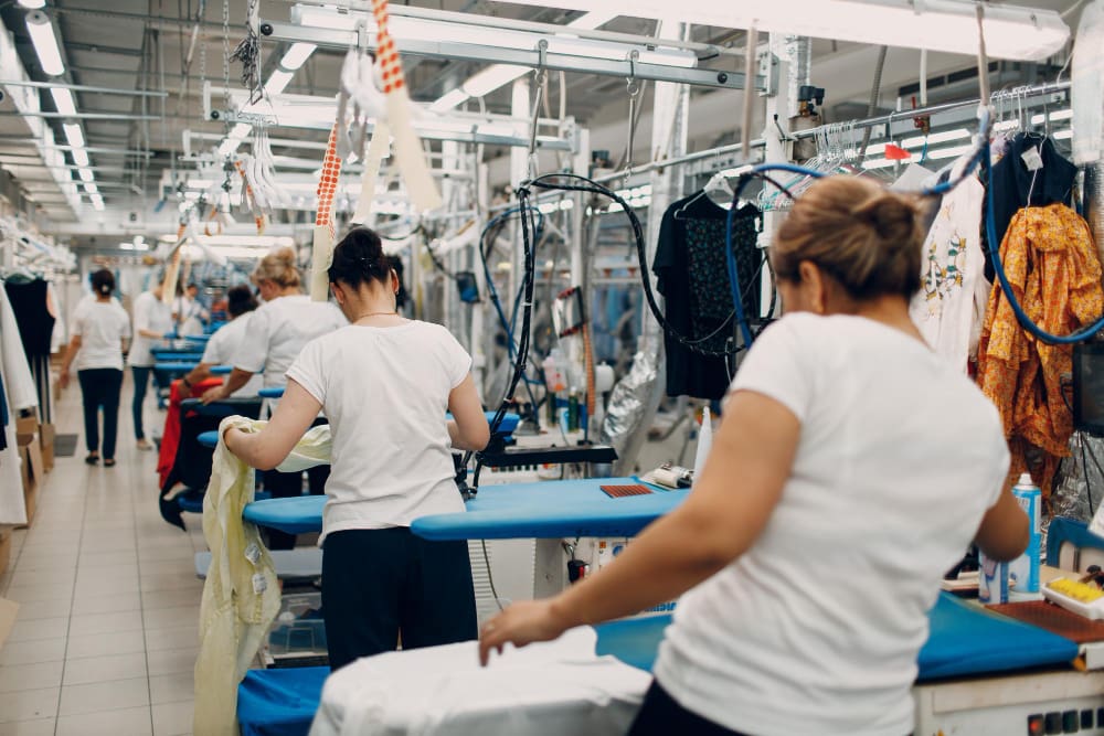 Workers in a garment factory use ironing boards and steam presses to prepare clothing items in a well-lit, busy production line.