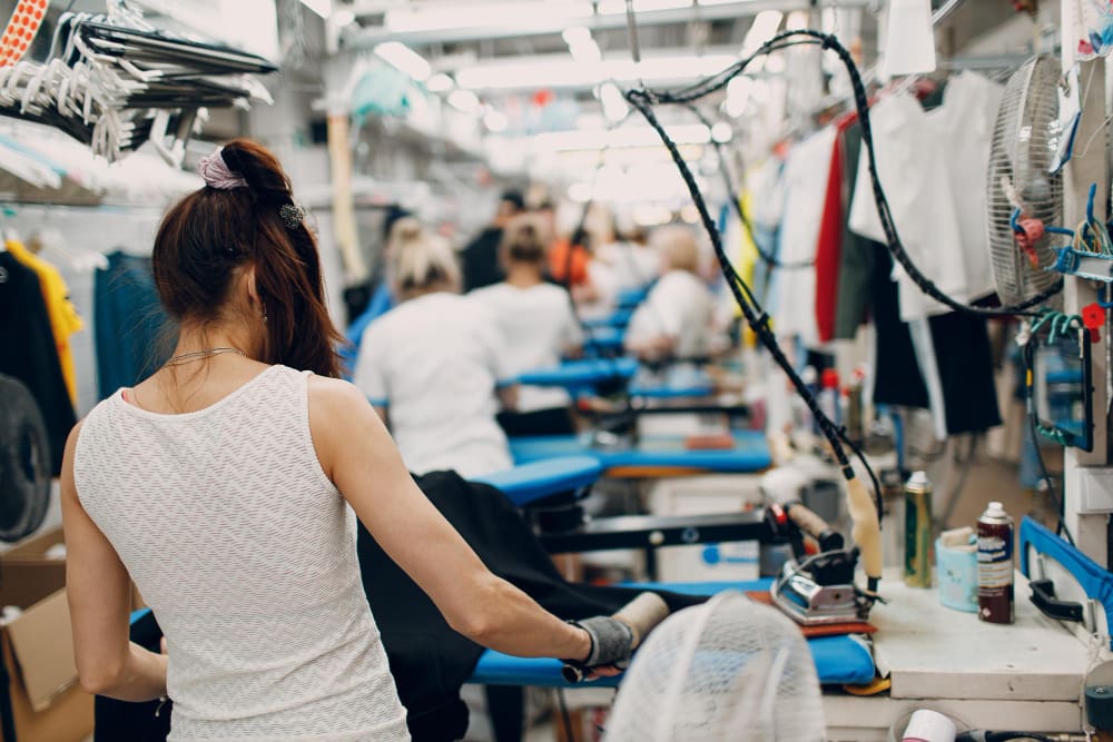 A group of workers in a garment factory sit at workstations ironing clothes, with clothing and equipment visible around them.