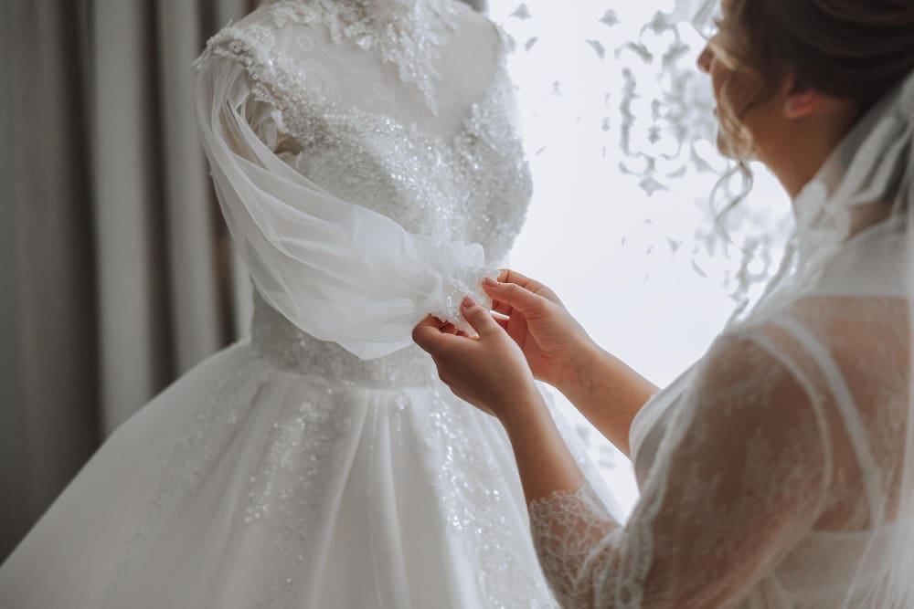 A woman in a lace robe adjusts the sleeve of a white, beaded wedding dress displayed on a mannequin near a window.