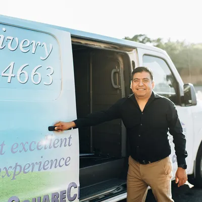 A man in a black shirt and beige pants stands by the open sliding door of a white delivery van, smiling at the camera.