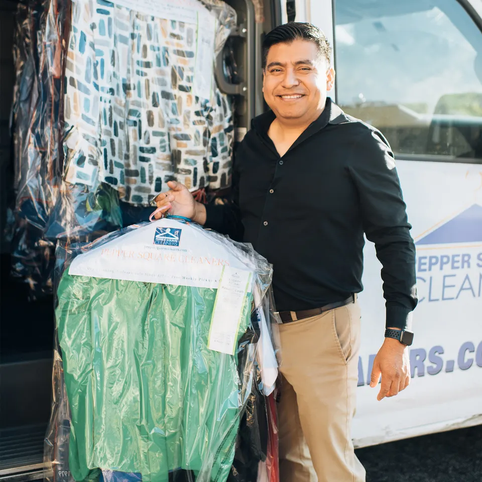 A man stands next to a van holding freshly cleaned clothes covered in plastic from a dry cleaning service.
