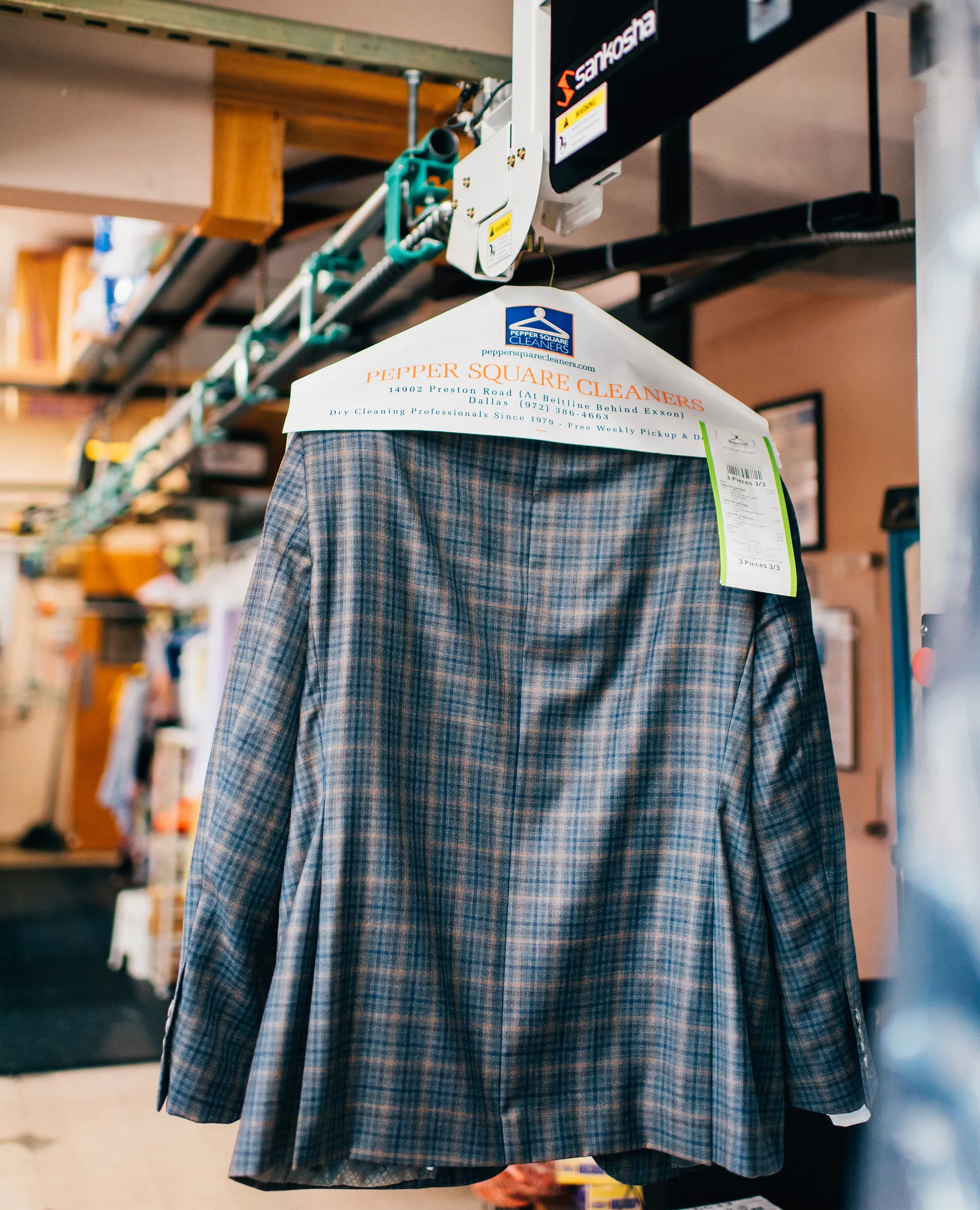 A plaid suit jacket hangs on a Peppy Square Cleaners hanger on a dry cleaning conveyor with a green tag attached.
