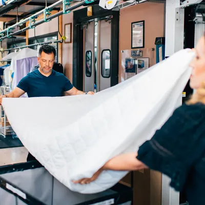 Two people work together to fold a large white sheet in a commercial laundry facility.