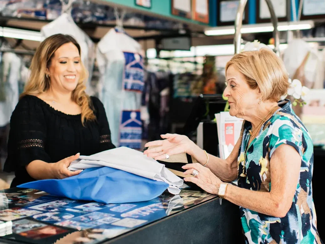 Two women stand at a dry cleaning counter; one hands folded clothes to the other, who is engaging in conversation.
