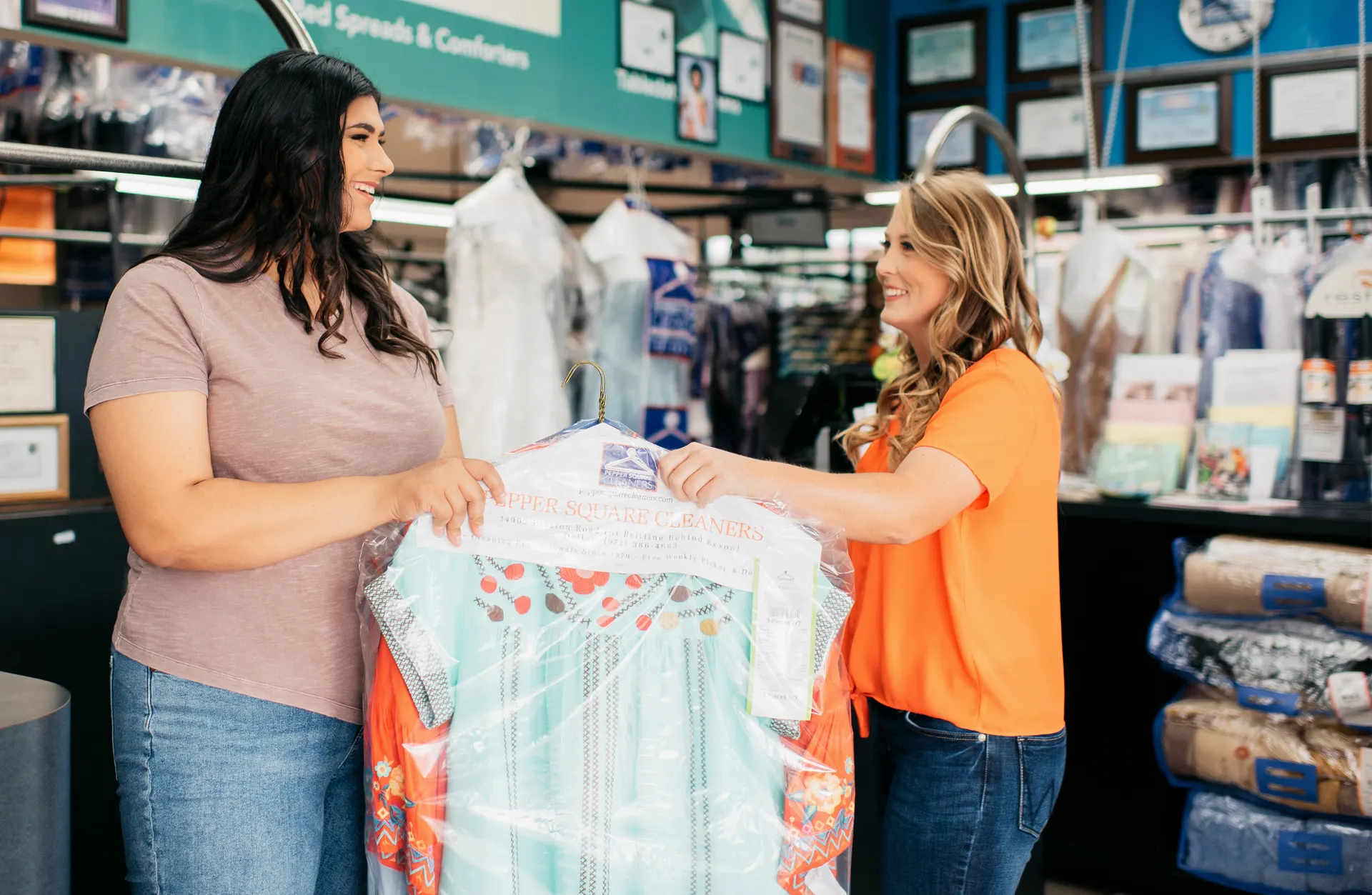 Two women stand in a dry cleaning shop, smiling and holding freshly cleaned clothes wrapped in plastic. Clothing racks and certificates are visible in the background.