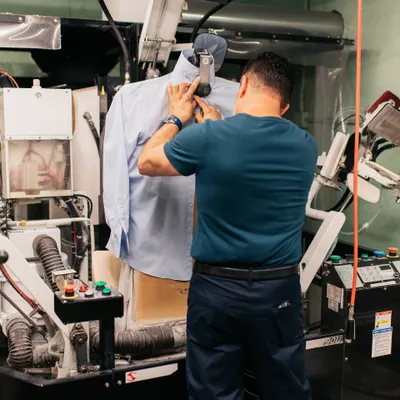 A man operates a machine that presses a button-down shirt on a mannequin in an industrial laundry or garment facility.