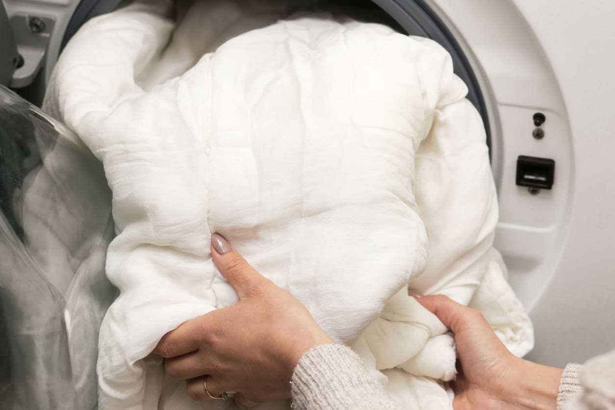 A person loads a large white comforter into a front-loading washing machine.