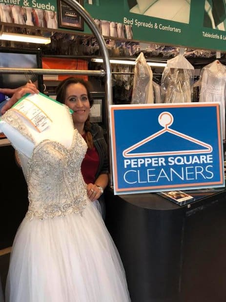 A woman stands beside a wedding dress on a mannequin at a counter next to a sign that reads "Pepper Square Cleaners.