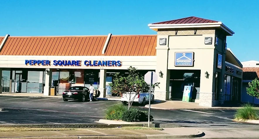A small shopping center with a storefront labeled "Pepper Square Cleaners" and a car parked in front on a sunny day.
