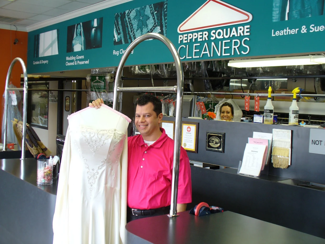 A man in a pink shirt holds a white wedding dress at the counter of a dry cleaning shop called Pepper Square Cleaners.