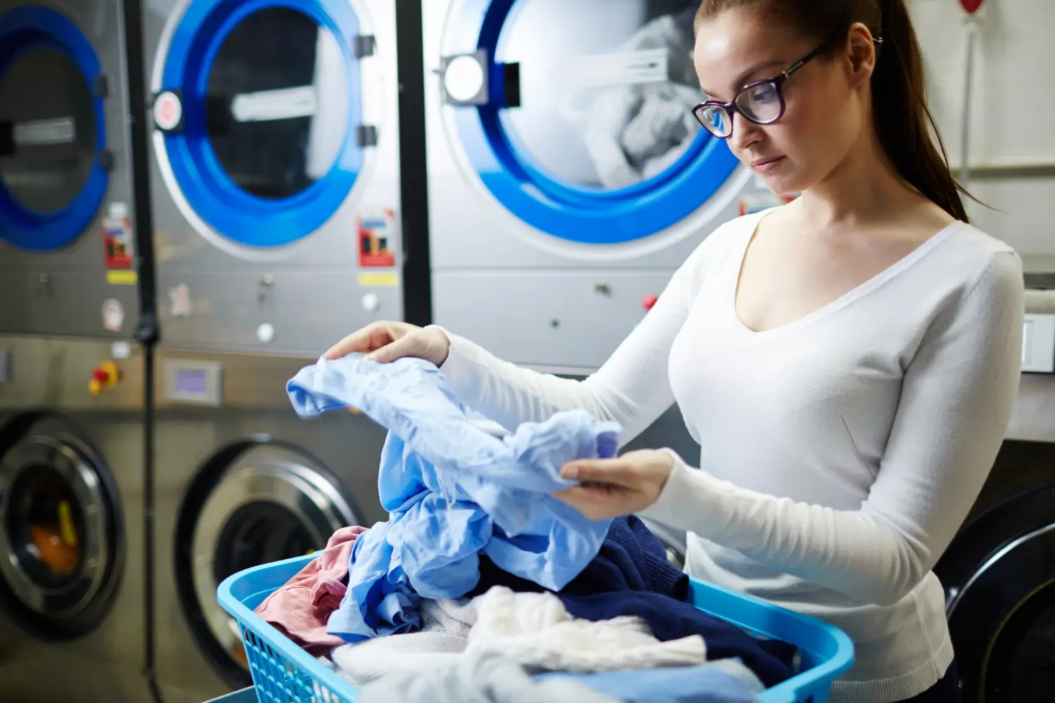 Woman wearing glasses sorts laundry from a blue basket in front of industrial washing machines at a laundromat.