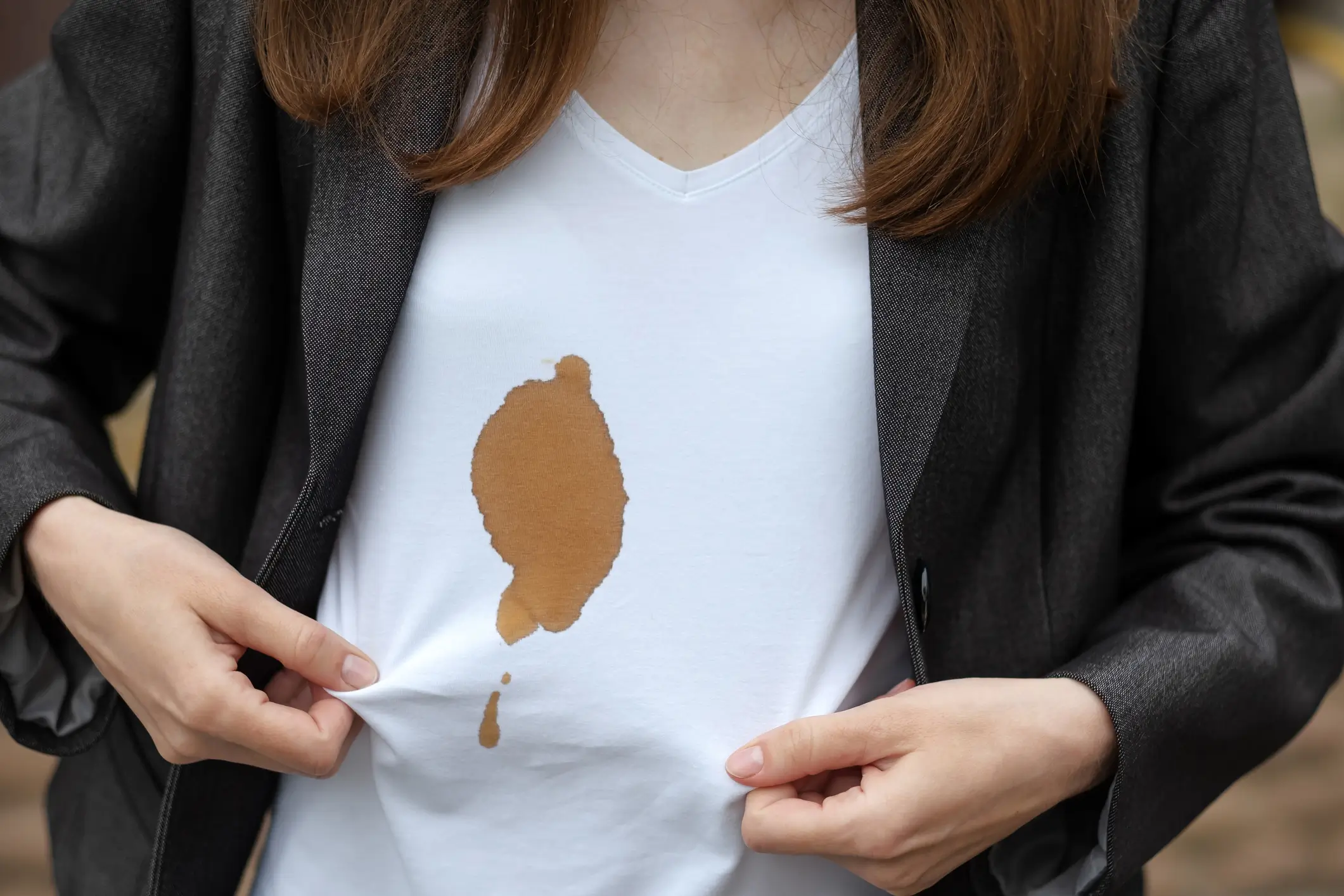 A person in a blazer holds up their white t-shirt, which has a large brown stain on the front.